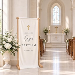Pode incluir: Uma faixa vertical de tecido branco num suporte de madeira clara. A faixa apresenta um ícone de cruz dourada no topo, seguido pelo texto "WELCOME TO", "Enzo's", "BAPTISM" e a data "OCTOBER 18, 2026" em letras pretas.