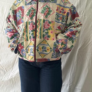 Peut inclure: Veste à motifs floraux multicolores. La veste est ornée d'un motif patchwork avec des fleurs roses, rouges, jaunes et bleues. Elle est dotée d'une fermeture éclair et d'un col. Portée avec un jean bleu foncé.
