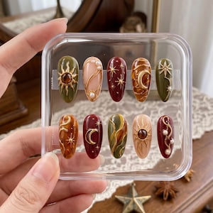 May include: A set of ten press-on nails in a clear plastic case, featuring almond-shaped tips with celestial designs. The nails display olive green, deep red, amber, and nude tones, accented with gold-colored sun, moon, and star motifs and pearls.