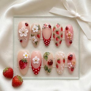 May include: A set of ten artificial nails with a light pink base, featuring various designs including strawberries, flowers, ladybugs, and red polka dots. Some nails have 3D floral accents. The nails are displayed on a clear glass surface.