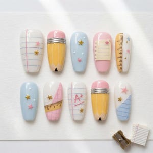 May include: A set of ten press-on nails featuring school-themed designs including notebook paper with blue and pink lines, yellow pencils with silver ferrules, light blue backgrounds with gold stars, a ruler, and a paper with an A+ grade written in red.