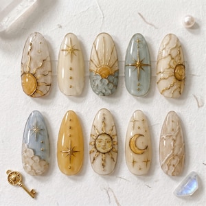 May include: A set of ten almond-shaped press-on nails featuring celestial designs. The nails display gold-toned suns, moons, stars, and clouds against cream, yellow, and light blue backgrounds. Some nails have raised textures and metallic accents.