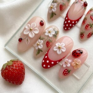 May include: A collection of almond-shaped artificial nails with a light pink base, adorned with white flowers, red strawberries, and ladybug designs. Some nails feature a red and white polka dot pattern. A fresh strawberry is in the foreground.