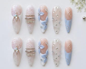 May include: A set of ten almond-shaped press-on nails featuring a variety of designs including white lace patterns, silver metallic rings, light blue floral accents, clear rhinestones, and silver swirl details on a nude and white base.