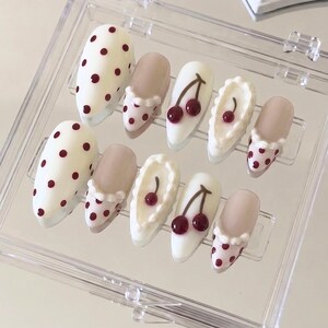 May include: A set of ten artificial nails in a clear plastic case. The nails feature various designs, including white with maroon polka dots, nude with a ruffled edge, and white with cherry designs. The nails are almond-shaped.