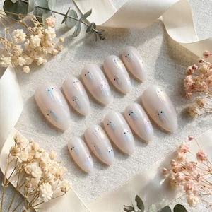 May include: A set of ten almond-shaped press-on nails with a glossy off-white base. Each nail features a small, iridescent ghost design with two tiny black dots for eyes, creating a subtle shimmering effect across the smooth surface of the nails.