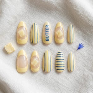May include: A set of ten almond-shaped press-on nails in pale yellow. Designs include vertical and horizontal blue stripes, a solid yellow nail with the word BUTTER in blue, and nails featuring a raised, three-dimensional butter pat accent.