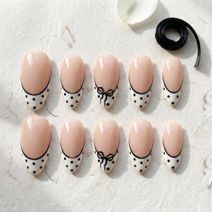 May include: A set of ten almond-shaped press-on nails featuring a nude base with white tips adorned with black polka dots. Two of the nails include a small black bow graphic centered on the tip. A small black adhesive strip is included in the set.