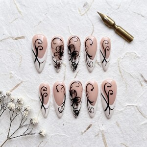 May include: A set of ten almond-shaped press-on nails featuring a nude base with white French tips and intricate black floral and scrollwork designs. The nails are arranged in two rows, accompanied by a metallic calligraphy pen nib.