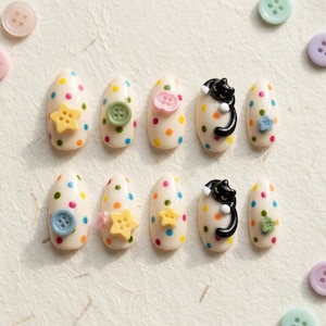 May include: A set of ten almond-shaped press-on nails with a cream base and multicolored polka dots. Each nail features a three-dimensional embellishment, including yellow stars, various colored buttons, small flowers, and a black cat figure.