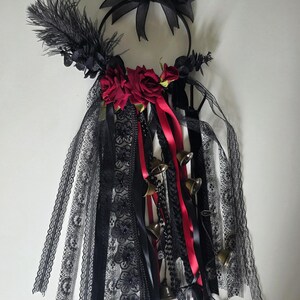 May include: A circular hoop wall hanging featuring a black mesh bow at the top, adorned with a black feather, two deep red fabric roses, and a cascading arrangement of black lace ribbons, red satin ribbons, and small metallic brass-toned bells.