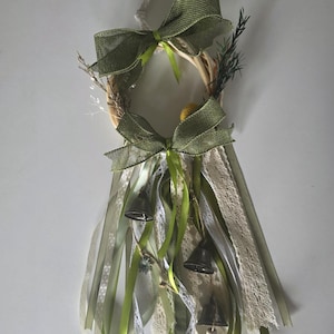 May include: A decorative wall hanging featuring a circular wooden hoop adorned with two olive green burlap bows, dried foliage, and multiple hanging ribbons in shades of green and white lace. Three small metal bells are suspended from the ribbons.