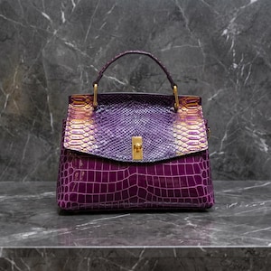 May include: A purple handbag with a textured crocodile pattern on the lower section and a snakeskin pattern on the upper flap. The bag has a gold-coloured clasp and handle attachments.