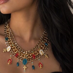 May include: Gold-toned layered necklace with religious pendants. The necklace features multiple chains of varying thicknesses. Pendants include crosses, oval medallions with religious figures, and colorful beads. The necklace is worn against a dark background.