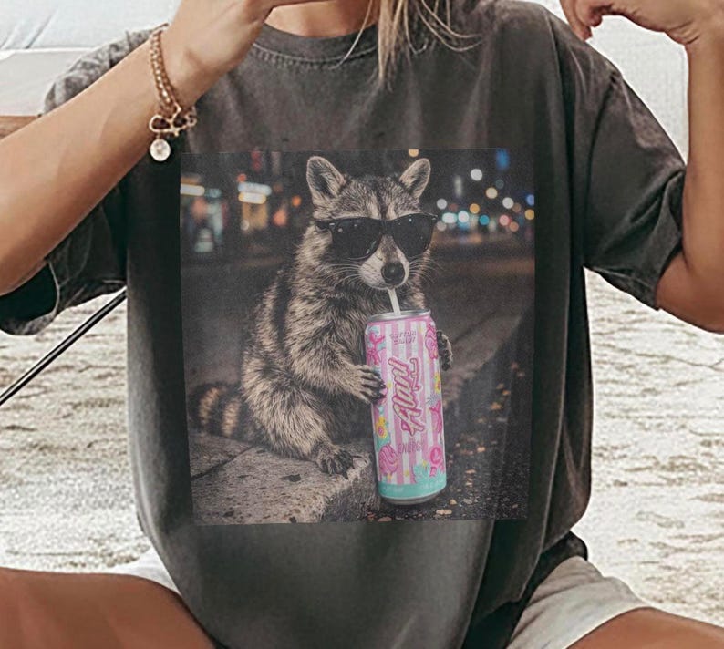 Pu&ograve; includere: Una t-shirt grigio antracite con una stampa grafica di un procione che indossa occhiali da sole neri e tiene in mano una lattina a righe rosa e bianche con il testo Alani stampato sul davanti, su uno sfondo sfocato di una strada cittadina.