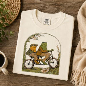May include: Cream-colored t-shirt featuring a whimsical illustration of two frogs riding a tandem bicycle. The frogs are dressed in vintage clothing, with one wearing a cap. The design is framed by an archway of foliage.