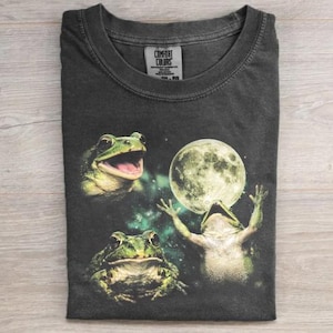 May include: Dark gray Comfort Colors t-shirt featuring a whimsical design of three green frogs. Two frogs are positioned on the left, and one frog is reaching for a full moon. The shirt has a crew neck and a tag in the upper center.