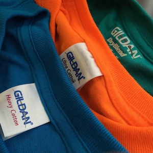 Pu&ograve; includere: Tre magliette in blu, arancione e verde sono sovrapposte. Ogni maglietta presenta un'etichetta interna bianca sul colletto con il logo blu Gildan. Le etichette specificano rispettivamente i tipi di tessuto Heavy Cotton, Ultra Cotton e DryBlend.