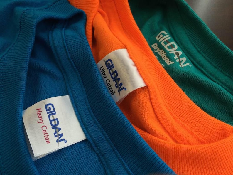 Pu&ograve; includere: Tre magliette in blu, arancione e verde sono sovrapposte. Ogni maglietta presenta un'etichetta interna bianca sul colletto con il logo blu Gildan. Le etichette specificano rispettivamente i tipi di tessuto Heavy Cotton, Ultra Cotton e DryBlend.