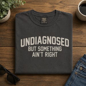 May include: A charcoal gray short-sleeve t-shirt featuring the text "UNDIAGNOSED BUT SOMETHING AIN'T RIGHT" in a light beige, arched, sans-serif font. The shirt has a crew neckline and a Comfort Colors brand label on the inside collar.