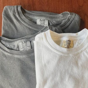 Pu&ograve; includere: Tre magliette a girocollo impilate, composte da una maglietta bianca e due magliette grigie. Ogni maglietta presenta un'etichetta del marchio Comfort Colors cucita all'interno del colletto, con il testo Comfort Colors sulle etichette.