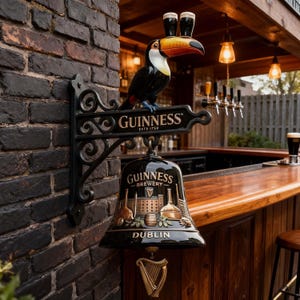 May include: A black Guinness Brewery sign with a toucan holding two glasses of stout. The sign is mounted on a brick wall and includes the text "Guinness Estd 1759" and "Dublin". A gold harp hangs below the bell.