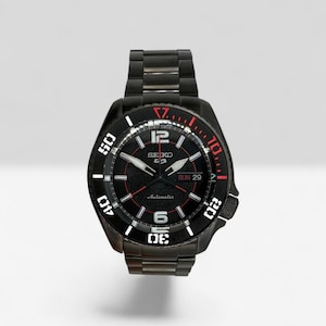 Puede incluir: Un reloj de pulsera automático Seiko 5 negro con correa de eslabones de acero inoxidable. La esfera negra presenta marcadores de hora blancos, números arábigos en el 6 y el 12, ventana de día y fecha a las 3 y detalles rojos en el bisel.