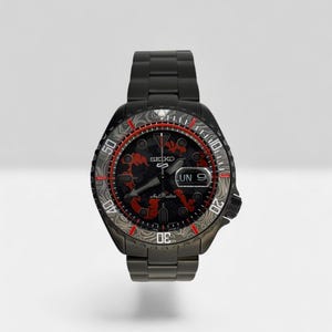 Peut inclure: Une montre automatique Seiko 5 noire avec un bracelet à maillons en métal. Le cadran présente un motif noir et rouge, des aiguilles luminescentes et un guichet jour-date à trois heures. La lunette rotative arbore un motif topographique gris.