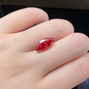 May include: A marquise-cut gemstone with a vibrant red color and faceted surface. The stone features a pointed oval shape and displays internal light reflections throughout its structure. A small logo with the letters GV is visible below the stone.