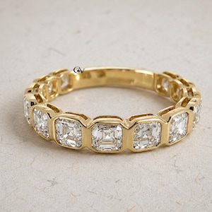 May include: A gold ring with a series of square-cut, clear gemstones. The gemstones are set in individual bezels, forming a continuous pattern around the ring. The ring is presented on a neutral, textured surface.