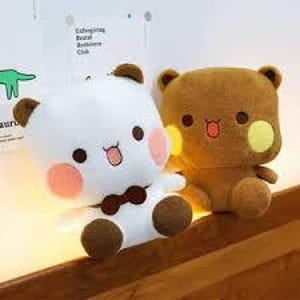 May include: Two plush bear toys. One is white with brown accents, a bow tie, and rosy cheeks. The other is brown with yellow cheek accents. Both have smiling faces and are sitting on a wooden surface.