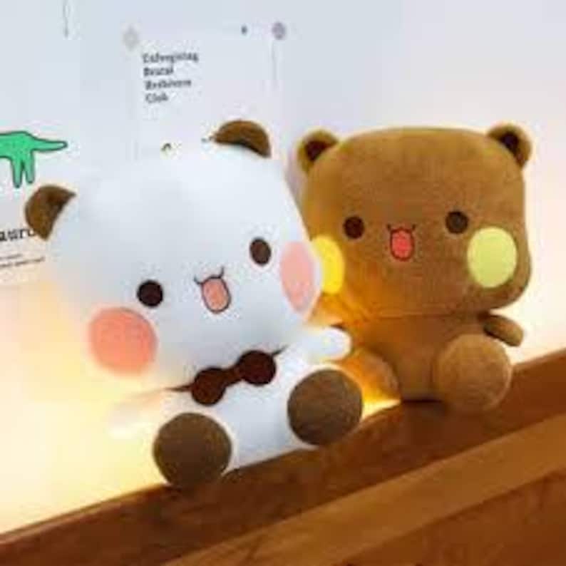 May include: Two plush bear toys. One is white with brown accents, a bow tie, and rosy cheeks. The other is brown with yellow cheek accents. Both have smiling faces and are sitting on a wooden surface.