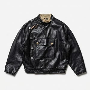 May include: A black leather jacket with a high collar and multiple pockets. The jacket features a button closure and a tan-colored lining visible at the collar. The jacket has a classic, vintage style.