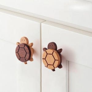 May include: Two turtle-shaped cabinet knobs attached to white cabinet doors. One knob features a light wood head and limbs with a dark wood shell, while the other knob features a dark wood head and limbs with a light wood shell.