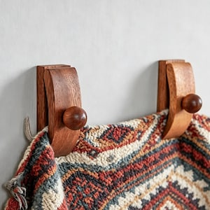 Può includere: Due ganci da parete in legno marrone medio, curvi e dotati di un pomello liscio e arrotondato ciascuno. I ganci sono montati su una parete e sostengono un tessuto spesso con un motivo geometrico nei toni del rosso, crema, blu e grigio scuro.