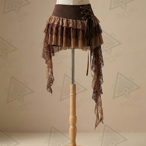 Op de afbeelding: Een bruine steampunk-stijl rok met meerdere lagen ruches en kant. De rok heeft een veterdetail in de taille en lange, vloeiende kanten panelen aan de zijkanten. De rok wordt getoond op een mannequin.