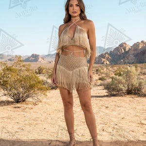 May include: A beige two-piece set consisting of a halter-style crop top and a matching mini skirt. Both pieces feature long, flowing fringe detailing and a textured, woven knit pattern. The top has thin straps that tie around the neck.