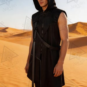 May include: Black hooded vest with an asymmetrical design. It has a front zip, leather strap details, and metal studs. The sleeveless vest has a long, flowing front panel. The background is a desert landscape.