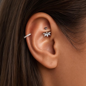May include: A silver-toned ear cuff featuring a row of small clear stones worn on the helix, paired with a rook piercing jewellery piece shaped like a fan with five marquise-cut clear stones and a small round metal backing disc.