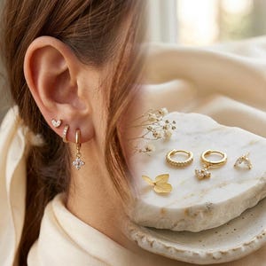 May include: A collection of gold earrings, including small hoops, a heart stud, and a flower-shaped dangle earring. The earrings are adorned with small, clear gemstones. The image is set against a soft, neutral background.