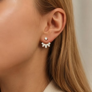 May include: A pair of gold-toned earrings with a stud and a decorative jacket. The stud features a single, round, clear gemstone. The jacket has a fan-like design with five marquise-cut gemstones. The earrings are worn on a person's ear.