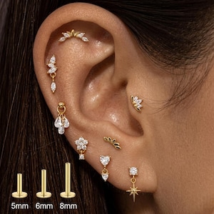 May include: A selection of gold earrings, including studs, drop earrings, and a curved design for the upper ear. Several earrings are adorned with clear gemstones. The image also shows three gold posts, measuring 5mm, 6mm, and 8mm in length.