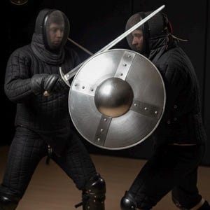 Peut inclure: Deux personnes en équipement de protection matelassé noir et masques d'escrime s'affrontent. L'une tient un bouclier rond en métal argenté avec un umbo central et des renforts rivetés, tandis que l'autre manie une longue épée en acier.