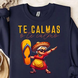 以下が含まれることがあります： ネイビーブルーのトレーナー。黄色と赤の文字で「TE CALMAS o te calmo」と書かれ、その下にソンブレロとサングラスを着用し、黄色いエプロン付きの赤いドレスを着た高齢者が、大きなオレンジ色のビーチサンダルを手に持っているグラフィックが描かれています。