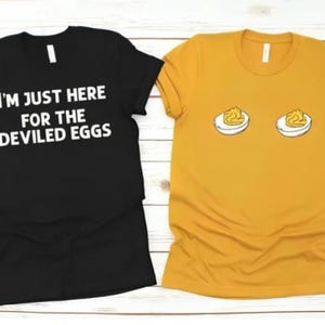 May include: Two t-shirts, one black and one mustard yellow, are displayed. The black shirt has the text "I'M JUST HERE FOR THE DEVILED EGGS" in white. The yellow shirt features a graphic of two deviled eggs.