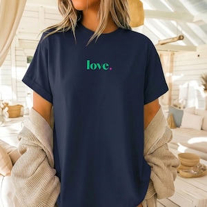 Könnte beinhalten: Ein marineblaues T-Shirt mit kurzen Ärmeln, auf dessen Brustmitte das Wort "love." in leuchtend türkiser Schrift gedruckt ist, gefolgt von einem kleinen pinken Punkt. Das Shirt hat einen Rundhalsausschnitt und eine lockere Passform mit umgekrempelten Ärmeln.