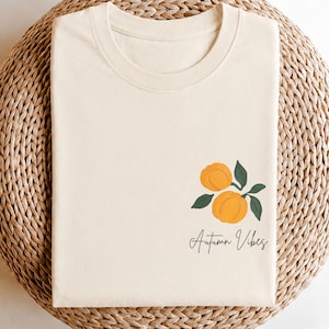 Könnte beinhalten: Ein cremefarbenes T-Shirt mit Rundhalsausschnitt und einer Grafik von zwei orangefarbenen Kürbissen mit dunkelgrünen Blättern auf der unteren rechten Seite. Darunter steht der Text "Autumn Vibes" in einer schwarzen Schreibschrift.