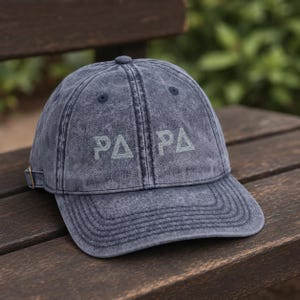 Puede incluir: Una gorra de béisbol de color azul oscuro con efecto lavado al ácido, construcción de seis paneles y visera curva. En la parte frontal presenta el texto bordado "PAPA" en una fuente estilizada de color gris claro con triángulos.