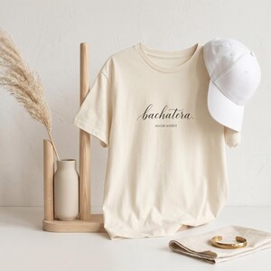 Puede incluir: Camiseta color crema con el texto "bachatera. social addict" en escritura negra. Una gorra de béisbol blanca descansa sobre la camisa. También se ve un jarrón beige con hierba de la pampa seca y una pulsera dorada.