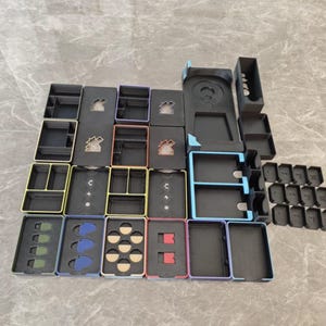 May include: A collection of black plastic board game organizer trays with various colored rims, including yellow, blue, red, and purple. The trays feature custom cutouts for game components, including circular, rectangular, and jagged shapes.